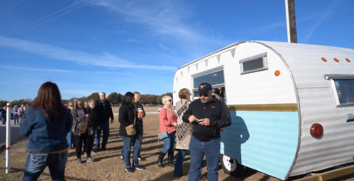 A crowd gathers around a vintage trailer on a sunny day, with people chatting and enjoying the outdoor atmosphere.