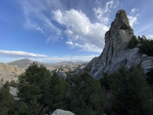 A rocky landscape with towering cliffs, green trees, and a blue sky with scattered clouds in the background.