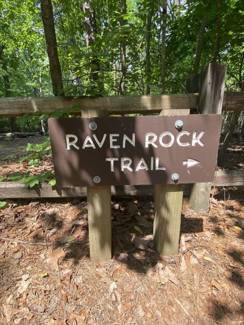 A wooden sign reading "Raven Rock Trail" with an arrow pointing to the right, surrounded by trees and foliage.