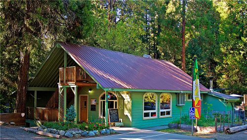 A green building with a metal roof surrounded by trees, featuring a sign and a welcoming entrance.