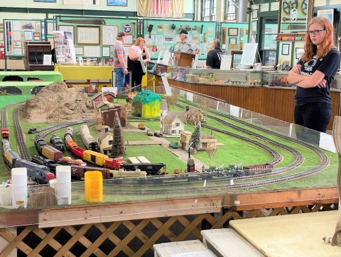 A model train display with tracks, buildings, and scenery, featuring a girl observing the setup in a museum-like setting.
