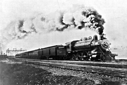 A vintage steam locomotive pulls passenger cars along a track, billowing smoke against a cloudy sky.