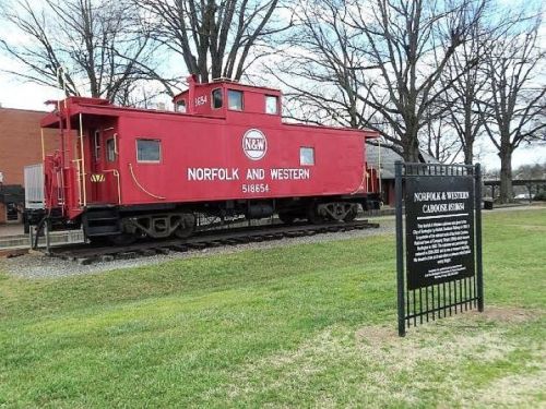 A red Norfolk and Western caboose on display next to a sign at a railway museum, surrounded by grass and trees.