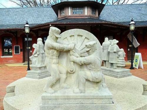 Stone sculpture depicting two workers operating machinery, set in front of a historic building with a slate roof.