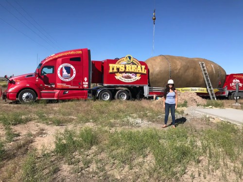 A woman stands beside a large potato-shaped truck in a grassy area under a clear blue sky.