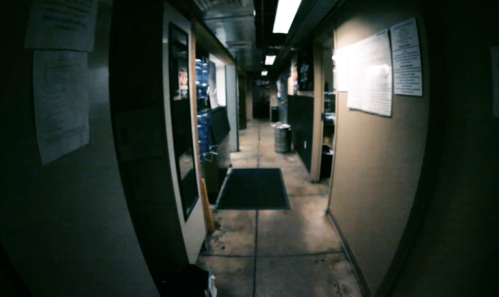 A dimly lit hallway with walls lined with papers and doors, featuring a concrete floor and overhead lights.