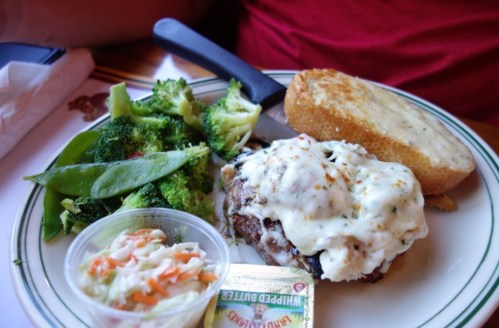 A plate with a burger topped with creamy sauce, garlic bread, steamed broccoli, snap peas, and coleslaw.