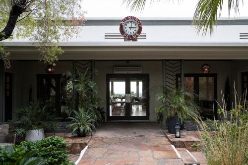 Entrance of a restaurant with a clock, surrounded by greenery and stone pathways.