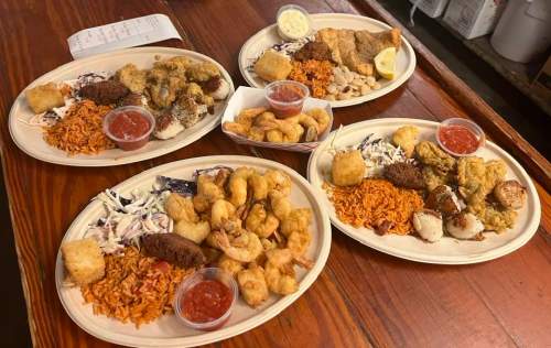 Four plates of fried seafood, coleslaw, and rice, with dipping sauces on a wooden table.