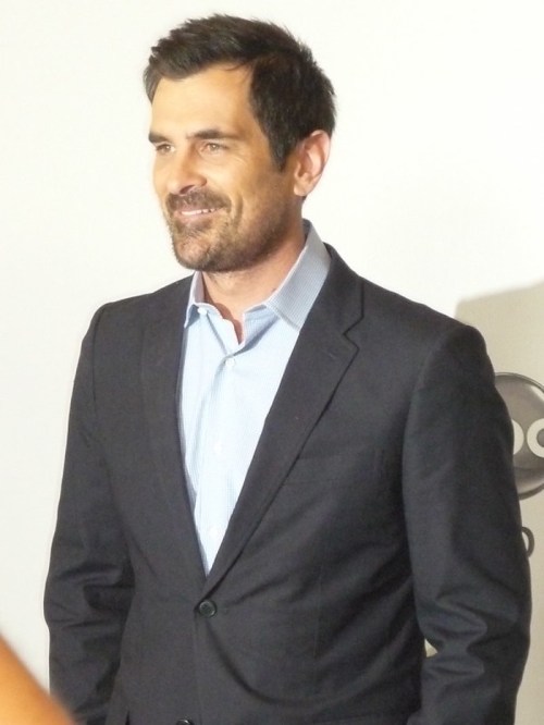 A man in a dark suit and light blue shirt smiles confidently at an event backdrop.