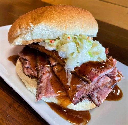 A hearty sandwich with layers of smoked meat, topped with coleslaw and drizzled with barbecue sauce on a soft bun.