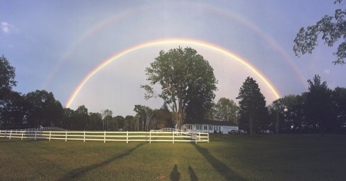 A double rainbow over a farm with a white fence.