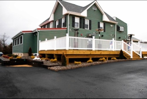 A green two-story house with a wooden deck and white railing, surrounded by landscaping and a paved driveway.