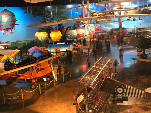 A museum display featuring various vintage aircraft, including biplanes and hot air balloons, in a spacious exhibit area.