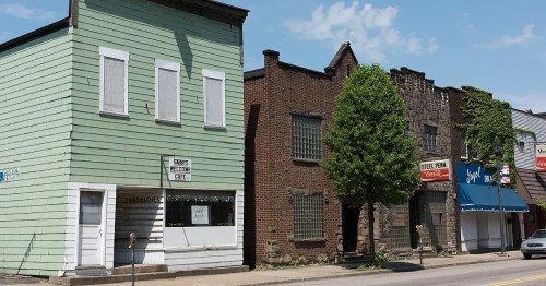 A row of buildings featuring a green structure, brick facades, and a large tree, under a clear blue sky.