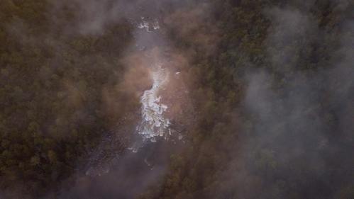 Aerial view of a river flowing through dense forest, partially obscured by mist and clouds.