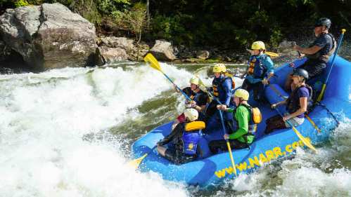 A group of people in a blue raft navigating whitewater rapids, wearing helmets and life jackets.