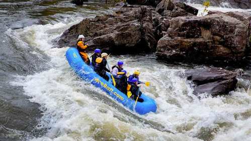 A group of four people in a blue raft navigating through whitewater rapids near rocky terrain.