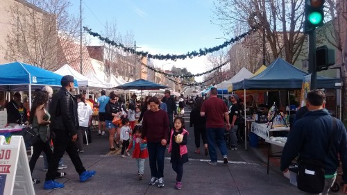 A bustling street market with colorful tents, people walking, and festive decorations overhead.