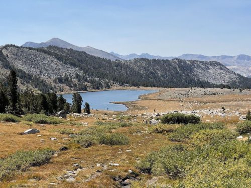 A serene landscape featuring a lake surrounded by mountains, trees, and rocky terrain under a clear blue sky.