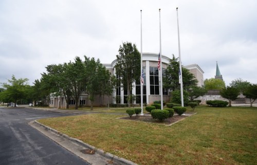 A modern building with trees and shrubs, featuring flagpoles in front and a cloudy sky above.