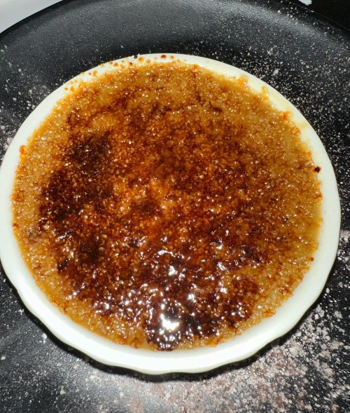 A close-up of a caramelized crème brûlée in a white dish, resting on a dark surface with a sprinkle of sugar.