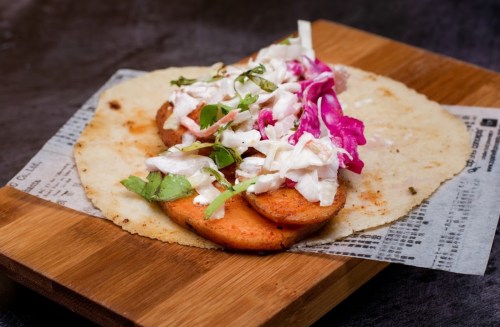 A wooden board holds a tortilla topped with sliced meat, cabbage, and herbs, garnished with pink onions.