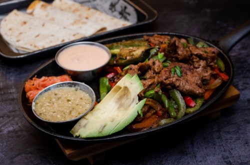 Sizzling fajitas with grilled meat, peppers, avocado, and sauces, served with flatbreads on a black platter.