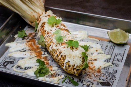 Grilled corn on the cob topped with cheese, cilantro, and drizzled with sauce, served on a metal tray with lime.