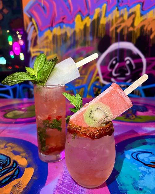 Two colorful cocktails with popsicles on top, garnished with mint, set against a vibrant, graffiti-style background.