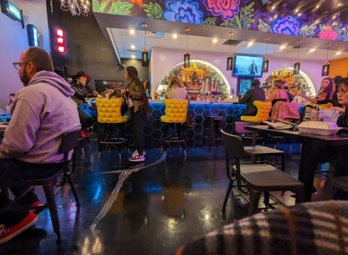 A vibrant restaurant interior with colorful decor, featuring patrons at tables and a bar with a variety of drinks.