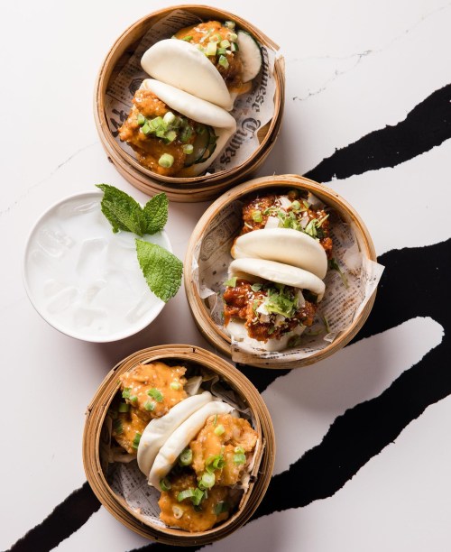 Three bamboo steamers filled with bao buns and fried chicken, garnished with green onions, alongside a glass of ice water.