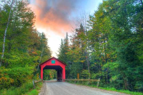 A red covered bridge surrounded by vibrant trees under a colorful sky at sunset.