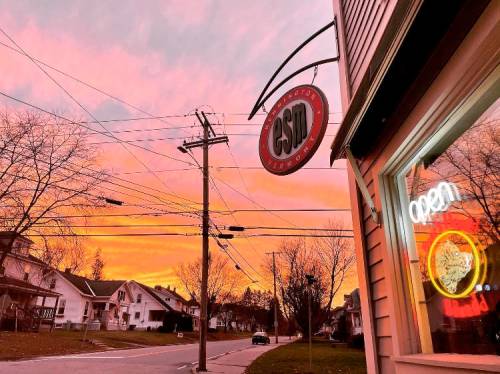 A vibrant sunset paints the sky pink and orange above a street with a shop sign and an "open" neon light.