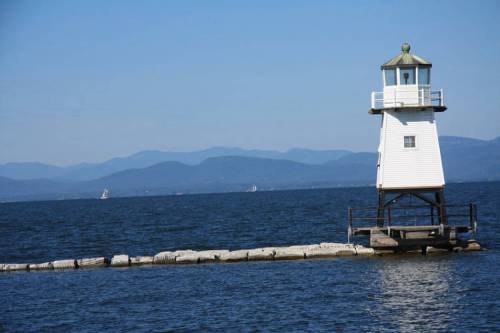 A white lighthouse stands on a rocky pier, surrounded by blue water and distant mountains under a clear sky.