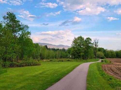A scenic path winds through lush greenery, with mountains in the distance under a blue sky with scattered clouds.