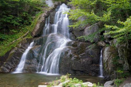 A serene waterfall cascading over rocks, surrounded by lush green trees and a calm pool at the base.