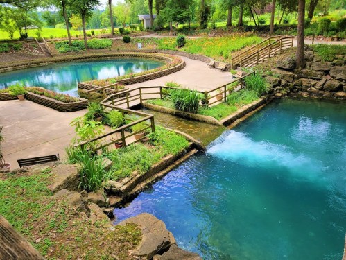 A serene park scene featuring two blue water bodies, wooden walkways, and lush greenery surrounding the area.