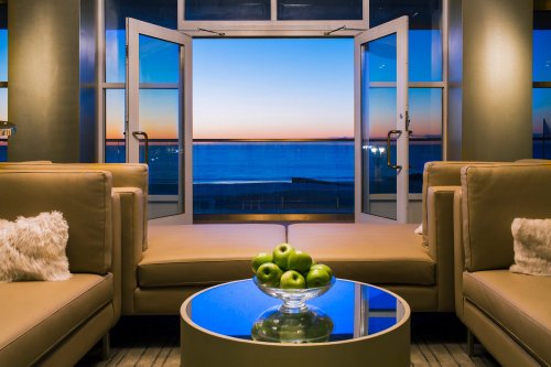 A cozy lounge with a view of the ocean at sunset, featuring a glass table with green apples and open double doors.