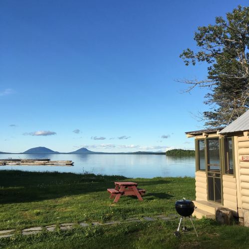 A serene lakeside scene featuring a cabin, picnic table, grill, and distant mountains under a clear blue sky.