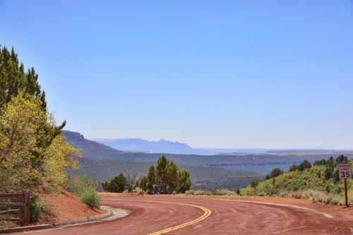 Winding road through a desert landscape with distant mountains and clear blue sky. Green shrubs line the roadside.