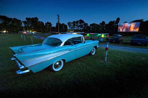 A vintage blue car parked at a drive-in theater, with a movie screen illuminated in the background at dusk.