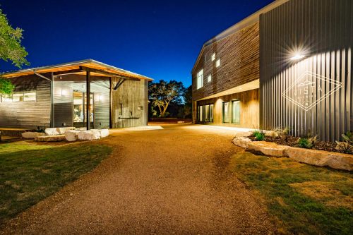 A modern building complex at night, featuring wooden and metal exteriors, with a gravel pathway and landscaped areas.