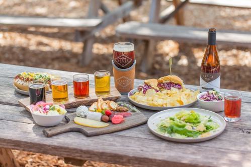 A variety of dishes and drinks on a wooden table, including burgers, chips, salads, and craft beers.