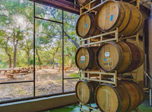 Wooden barrels stacked indoors, with a view of trees and picnic tables outside through large windows. Sunlight filters in.