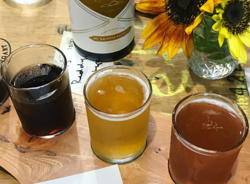 Three glasses of beer on a wooden board, with a bottle and a vase of sunflowers in the background.
