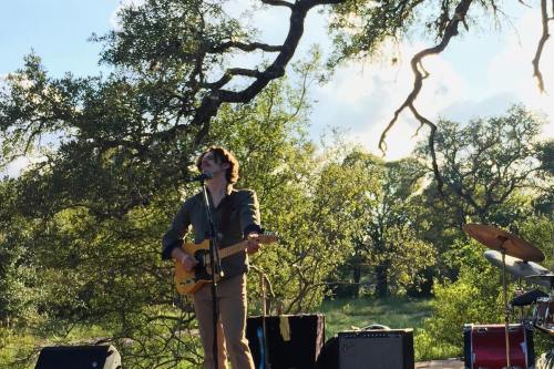 A musician sings and plays guitar on stage, surrounded by trees and greenery under a bright sky.