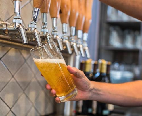 A hand pours beer from a tap into a glass, with several beer taps and wine bottles in the background.