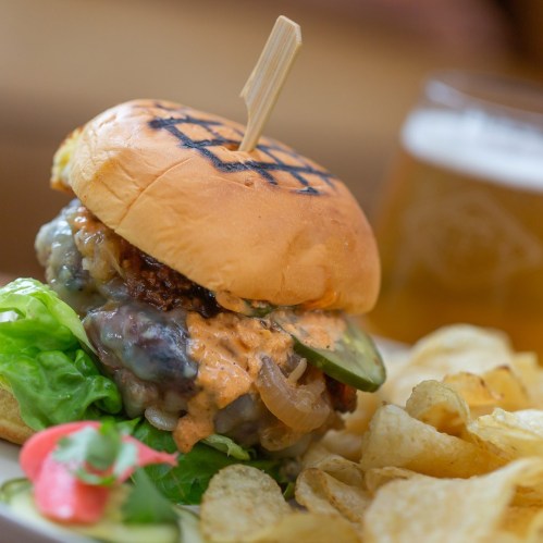 A juicy burger with lettuce, pickles, and sauce, served with chips and a glass of beer in the background.