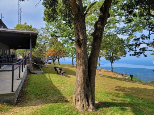 A scenic view from a grassy area with trees, benches, and a building, overlooking a distant landscape and blue sky.